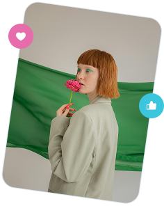 Woman with short red hair and green eye makeup holding a pink flower near her nose, standing in front of a green fabric backdrop.
